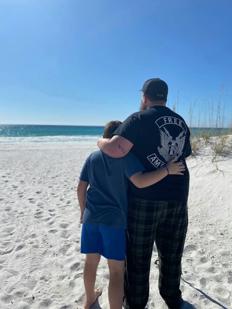 Bear with his arm around a boy on the beach, wearing a Free American eagle shirt and a BEAR cap